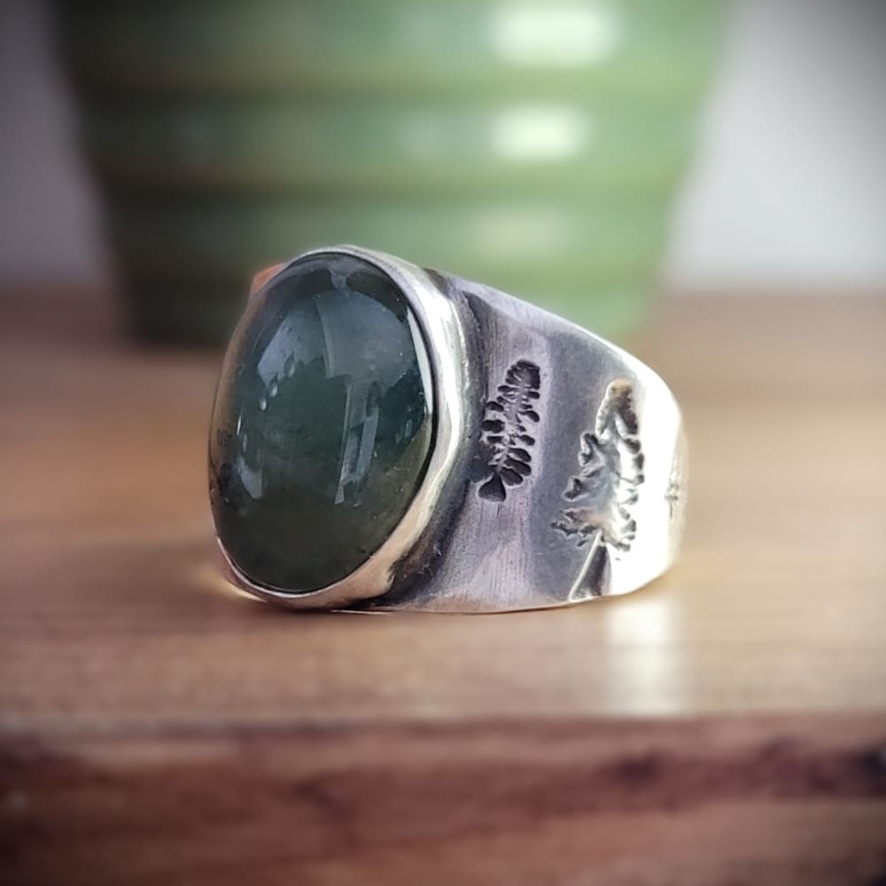 Moss agate cabochon set in wide sterling silver ring band featuring pine tree motifs.