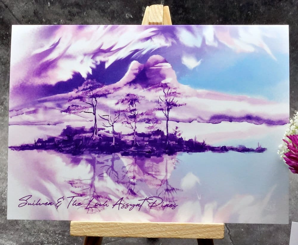 Larger image titled Loch Assynt Pines and Suilven showing an A6 size greetings card, blank inside, on a small easel depicting a view of old pine trees on islands on Loch Assynt, Sutherland in purple & blue watercolour.