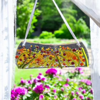 A vibrant fused glass suncatcher featuring orange, yellow, and green hues. The suncatcher has a curved shape and is hung by a white ribbon. Small pieces of glass are embedded in the design, creating a textured effect.