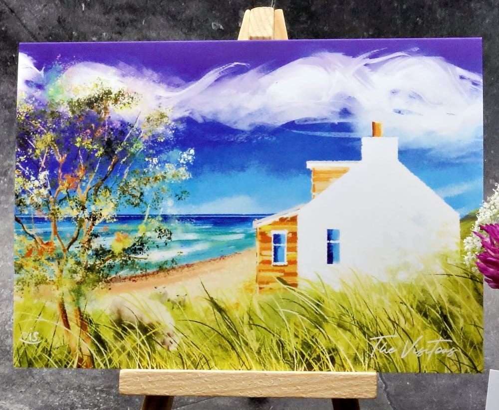 Larger image titled The Visitors showing an A6 size greetings card, blank inside, on a small easel depicting a view of a white cottage gable by the sea and sheep in the long grass on a breezy blue-sky day.