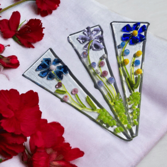 Three handcrafted fused glass plant stakes, each with a unique floral design, are displayed on a white cloth surrounded by vibrant red flowers. The stakes feature delicate flowers in blue, purple, and yellow tones, with intricate details in green glass, making them perfect for adding artistic flair to houseplants or garden decor.