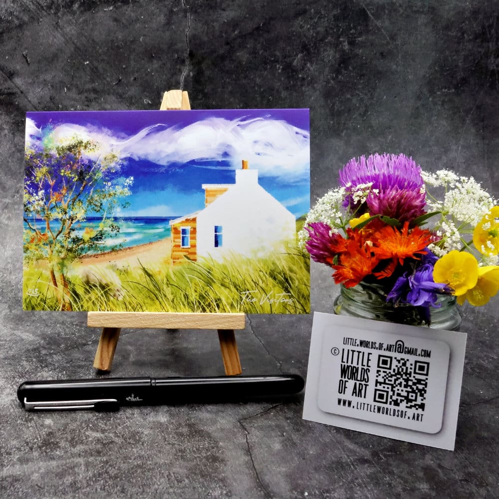 Image titled The Visitors showing an A6 size greetings card, blank inside, on a small easel depicting a view of a white cottage gable by the sea and sheep in the long grass on a breezy blue-sky day.