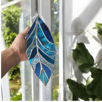 A hand holding a handmade stained glass suncatcher in the shape of a leaf or feather, made from various shades of blue glass. The suncatcher features intricate soldered lines and hangs by a white ribbon. In the background, there is a window with sunlight streaming through, highlighting the colourful glass, and a green plant is partially visible.