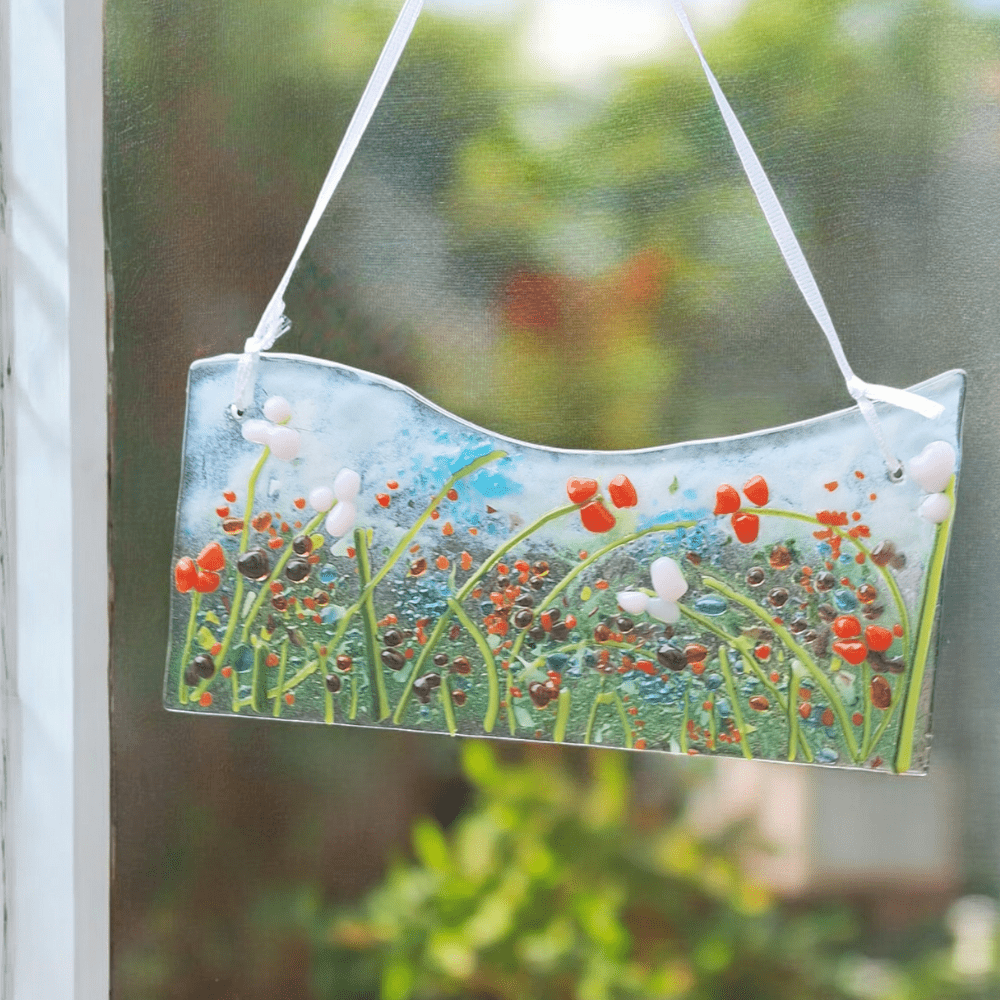 Handcrafted fused glass hanging picture featuring a vibrant floral design with red, white, and green elements, set against a blue and clear background. The glass art is suspended by a white satin ribbon and is displayed in a window with a soft, blurred outdoor scene in the background, highlighting the intricate details and bright colours of the fused glass decoration