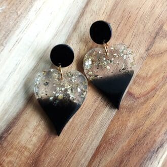 Drop Heart earrings made from black resin at the bottom of the heart and clear and gold flakes at the top. They are hung from flat black stud tops and are displayed on a wooden backdrop.