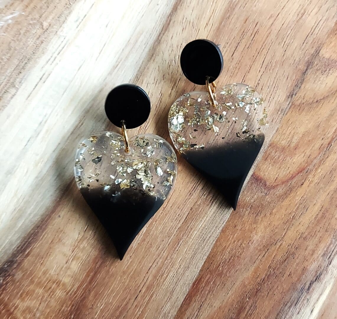 Drop Heart earrings made from black resin at the bottom of the heart and clear and gold flakes at the top. They are hung from flat black stud tops and are displayed on a wooden backdrop.