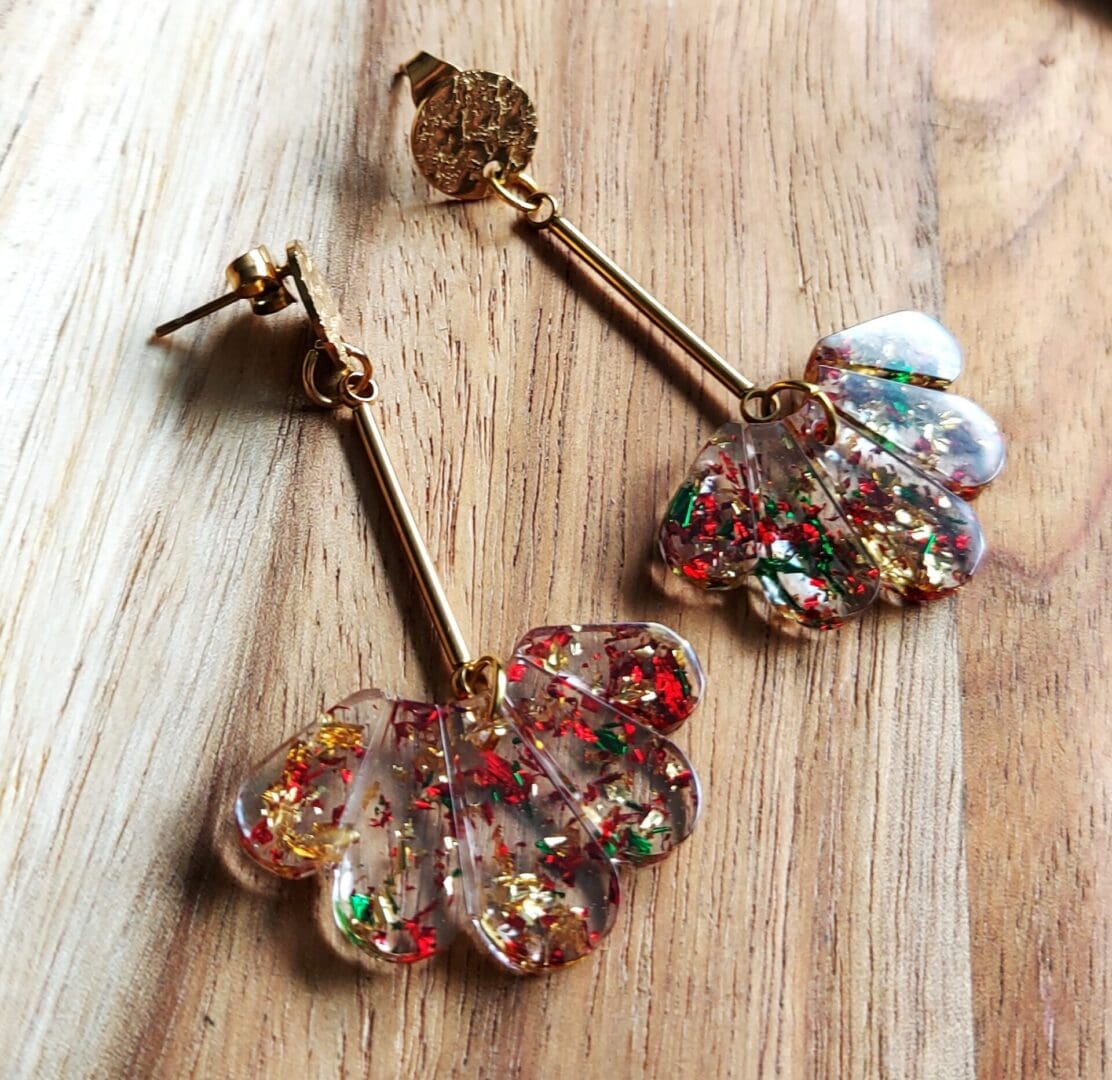 Art deco style fan drop earrings made from clear resin with red, gold and green foil flakes added. Hung from delicate gold wires which are fixed to textured gold tone stud tops. Displayed on a wooden backdrop.