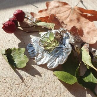 A solid silver hawthorn leaf and green sea glass pendant displayed in some autumn leaves