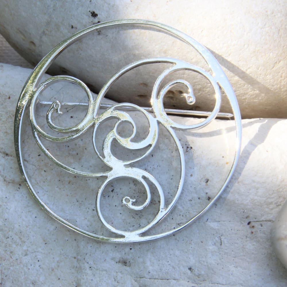 Sterling silver brooch in a round thick wire shape, inside the outer ring, three spirals interlocking in the middle forming another small circle. A duck face is at the end of each spiral. The brooch is set on white stones which serve as a background to the photograph.