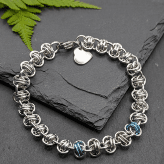 A chainmaille bracelet made with metal rings in silver, ice blue and pink in the style of a transgender pride flag as a section in the middle of the bracelet