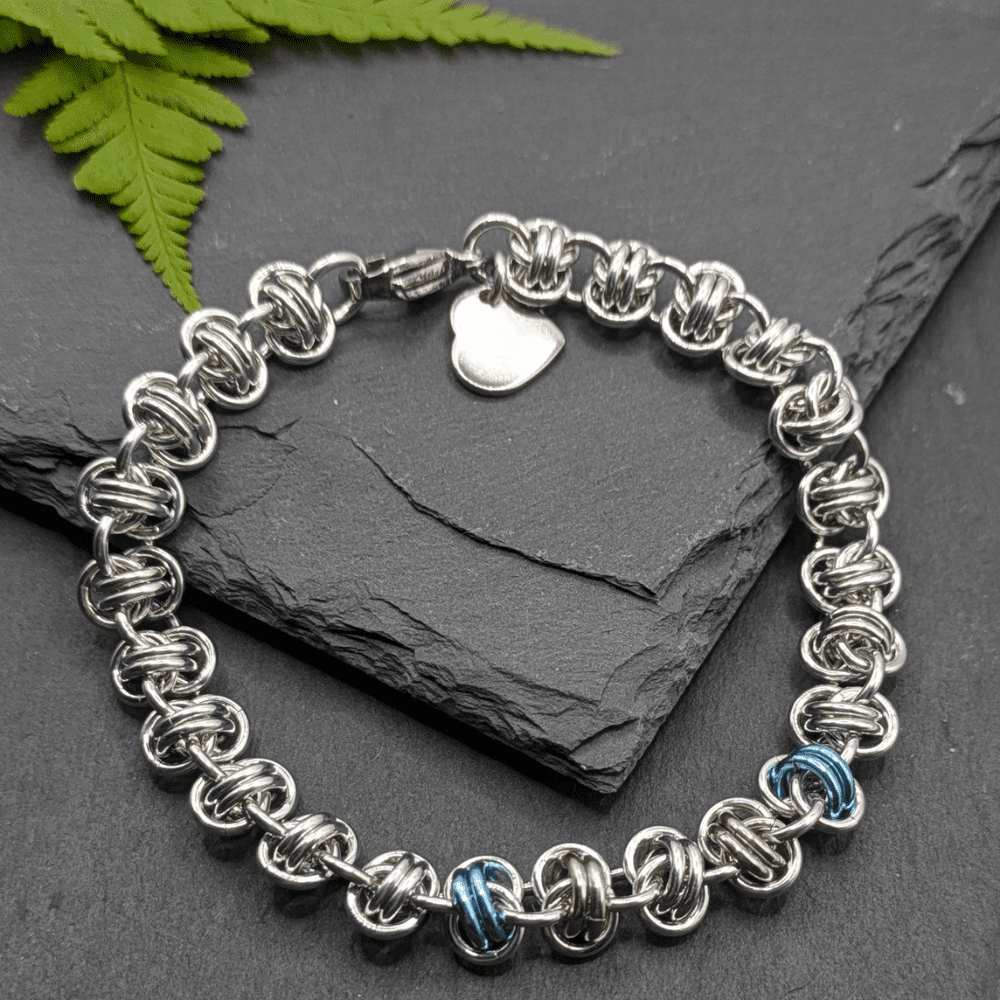 A chainmaille bracelet made with metal rings in silver, ice blue and pink in the style of a transgender pride flag as a section in the middle of the bracelet