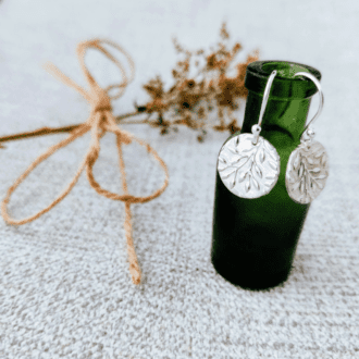 A pair of fine silver earrings with an olive branch design hung on a small green glass bottle with foliage in the background