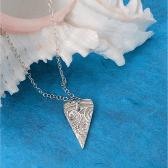 A fine silver ocean textured heart shaped pendant on a silver chain. Draped over an ornamental shell.