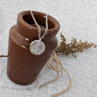 A silver pendant with an olive branch texture hung on a brown vintage jar with foliage in the background