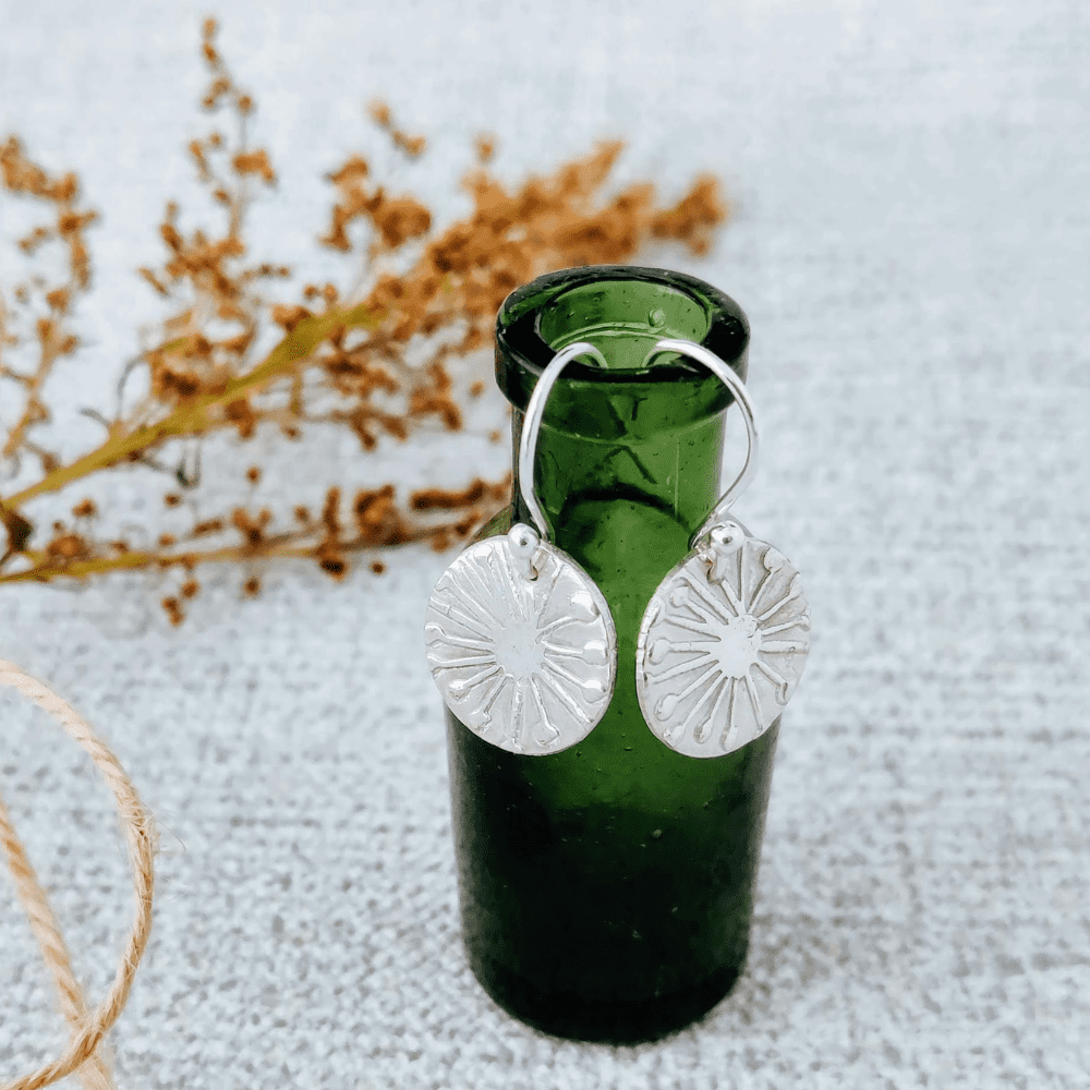 Fine silver drop earrings with a dandelion clock design hanging on a small green bottle. With foliage in the background