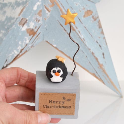 Handmade Merry Christmas decoration with grey wood base and clay penguin with gold star on wire.