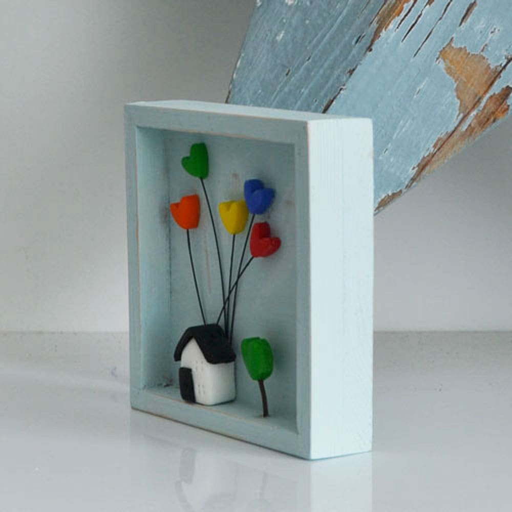 A miniature wooden frame with a rustic or distressed finish with a small clay house and rainbow hearts.