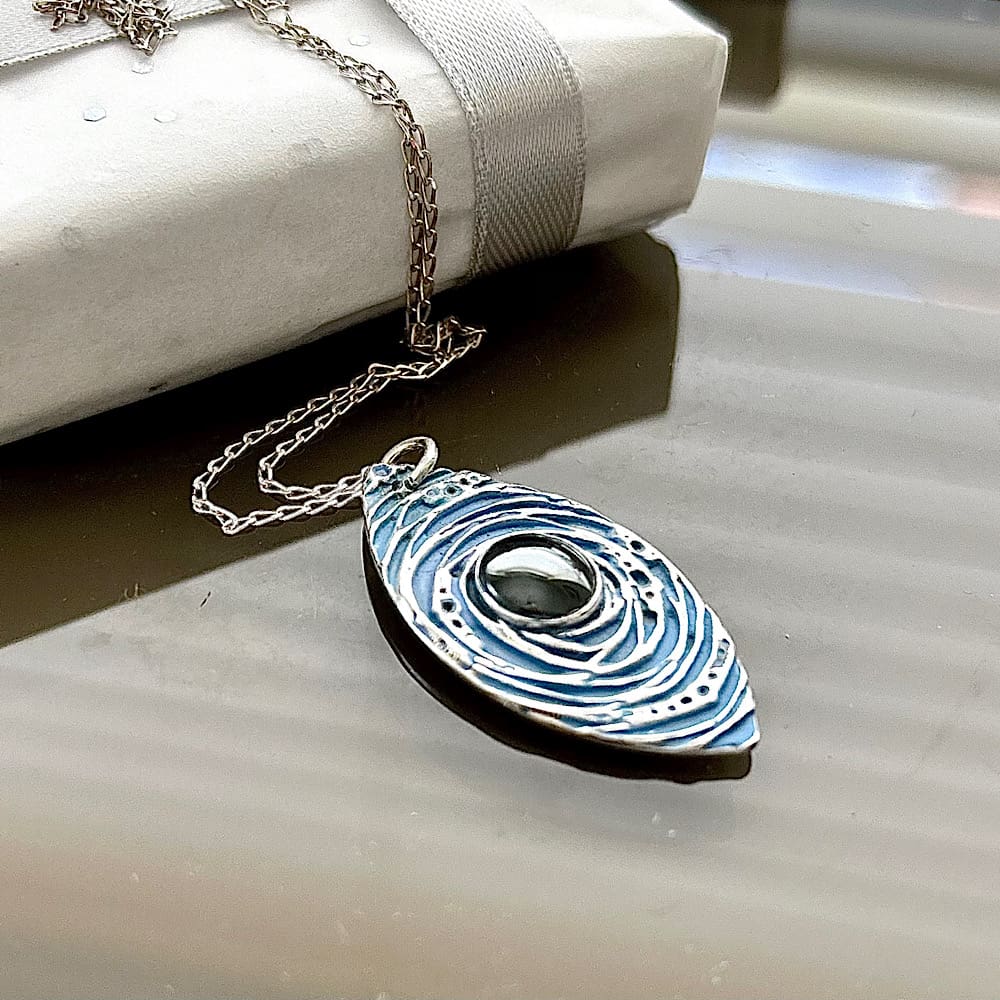 A silver pendant which is hand painted in pale blue with a hematite stone in the centre. With a sterling silver chain, displayed on a brown shiny surface.