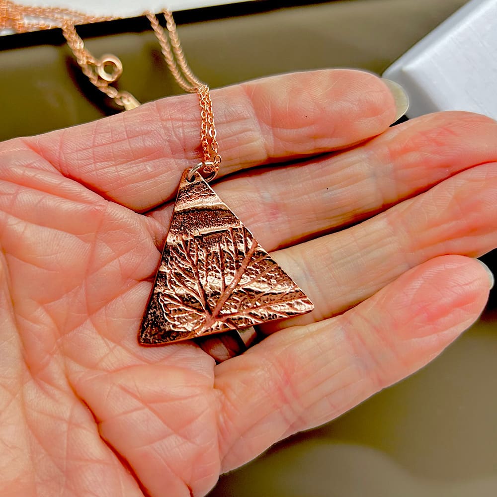 Copper pendant in a triangle shape with a leaf impression. Displayed on a white background showing a gift box.