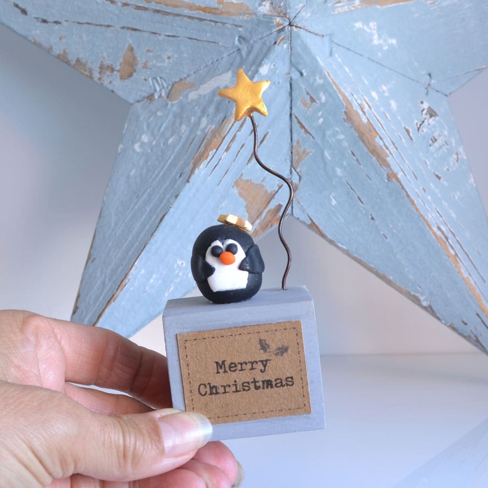 Handmade Merry Christmas decoration with grey wood base and clay penguin with gold star on wire.