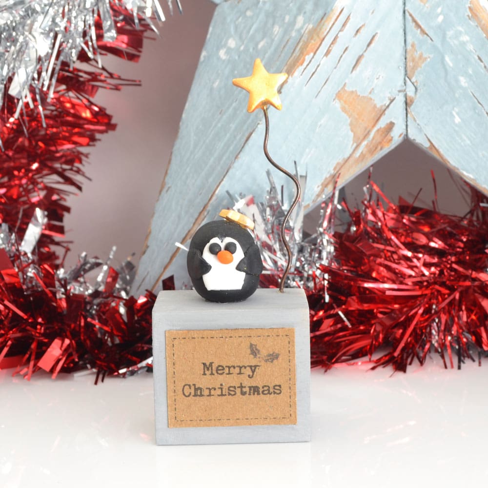 Handmade Merry Christmas decoration with grey wood base and clay penguin with gold star on wire.