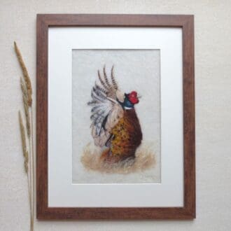 A handmade needle felted wool painting of a pheasant in a dark wood frame with a cream mount. The pheasant is facing right with wings raised.