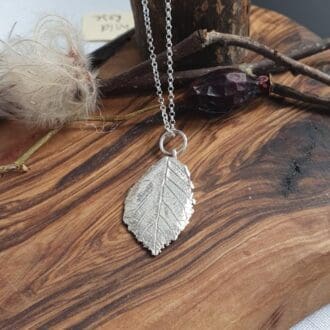 A solid silver wild rose leaf pendant necklace displayed on an olive board with autumn seeds