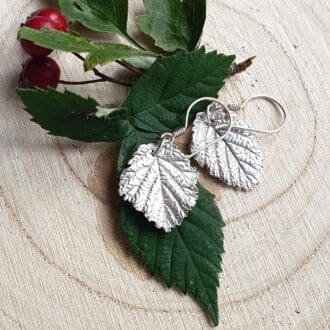 A pair of silver bramble leaf earrings displayed with bramble leaves on a wooden log slice.