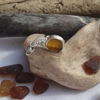 a chain bracelet with a tiny dark amber sea glass and silver charm displayed on aa piece of driftwood