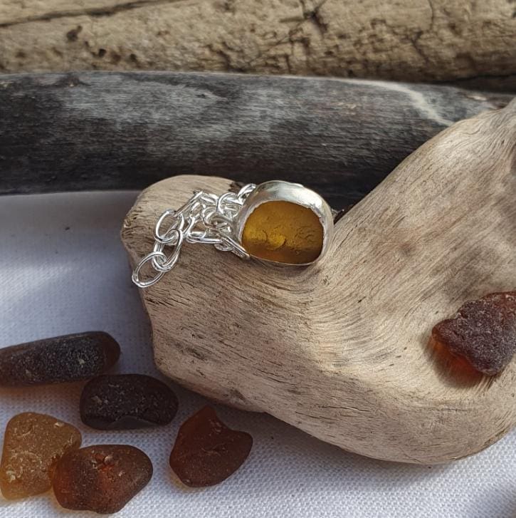 a chain bracelet with a tiny dark amber sea glass and silver charm displayed on aa piece of driftwood