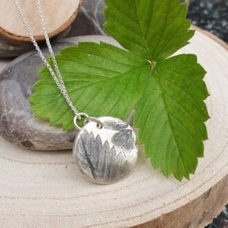 A solid silver circular pendant featuring a strawberry leaf anf flower imprint, displayed with strawberry leaves on wood