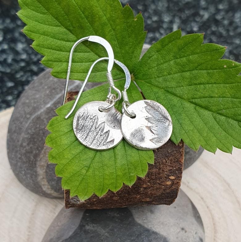 A pair of silver drop earrings imprinted with strawberry leaf and blossom displayed on some strawberry leaves