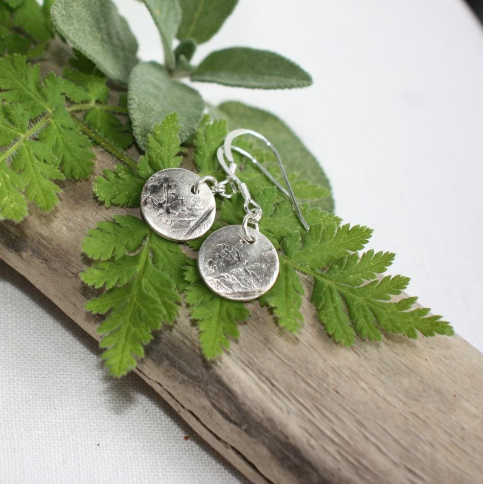 Silver drop disc shaped earrings with leafy imprints resting on herb leaves on a branch