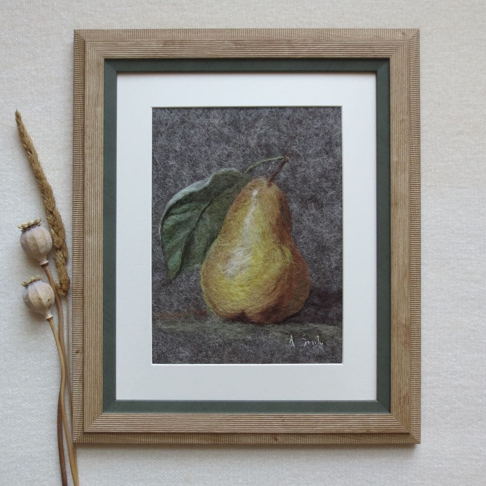 A wool felted picture of a gold coloured pear with a green leaf on a grey felt background. The picture has a cream mount and a pale wood frame with green inside edge.