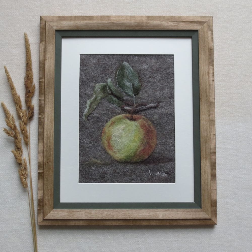 A handmade needle felted wool picture of an apple with leaves. The green and red apple is on a grey background and in a natural wood coloured frame with a green trim around the inside edge and a cream mount