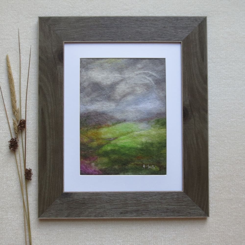 A wool felted picture of of a view across a Yorkshire Dales valley with the sun breaking through the clouds highlighting the fields. The picture has a white mount and is in a dark wood effect frame.