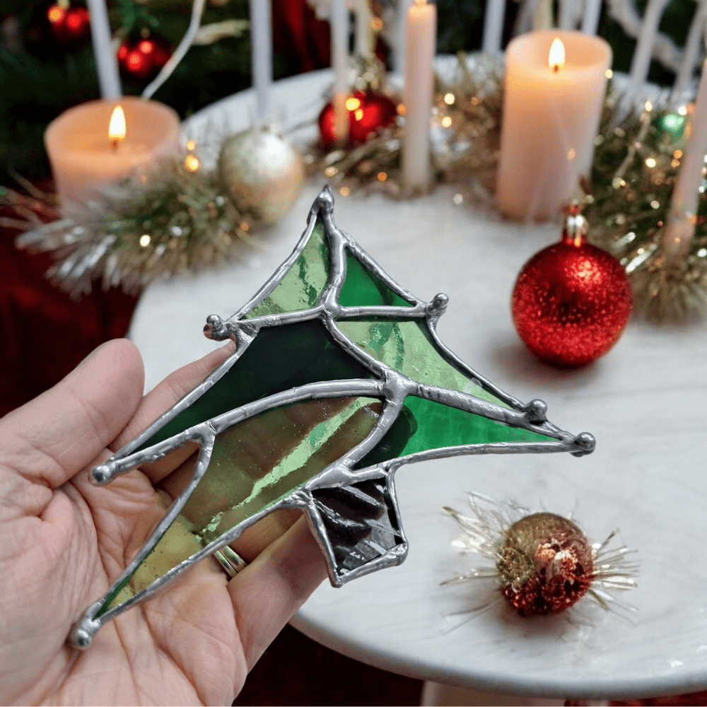 Handmade stained glass Christmas tree ornament with green glass and silver solder, perfect for holiday decor, Christmas gifts, and Secret Santa. Unique handcrafted ornament displayed in festive setting with candles and baubles