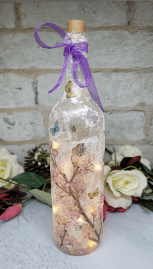 lit up standard wine bottle with flower butterfly design. Purple ribbon tied around the neck of a bottle