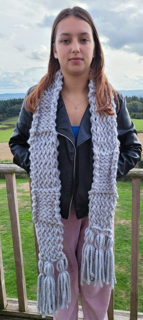 Long grey chunky knit scarf with large tassels seen around the neck of model. Scarf reaches knees