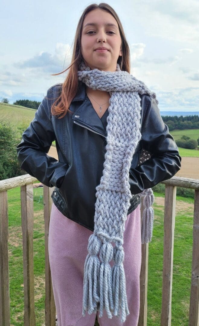 Grey chunky knit scarf with large tassels seen around the neck of model. Scarf reaches knees