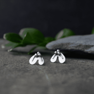 sterling silver stud earrings shaped like tiny mistletoe leaves with three tiny berries. Earrings rest on slate with foliage in background