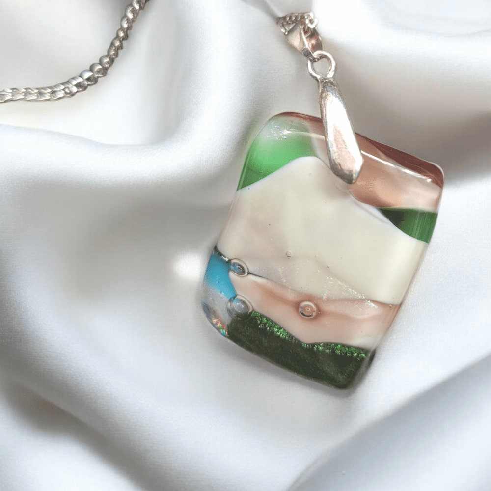 A handmade fused glass pendant featuring soft, layered colours of green, white, pink, and hints of dichroic glass. The rectangular pendant, with an abstract, landscape-inspired design, is attached to a silver-plated bail and chain. Perfect for a unique jewellery lover or as an anniversary gift.