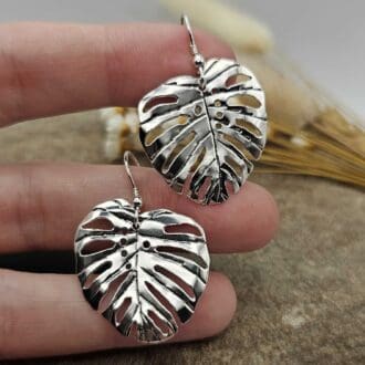 Pair of handcrafted recycled sterling silver monstera leaf drop earrings displayed hanging on jeweller's fingers