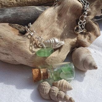 A chain bracelet with a tiny silver shell charm and a small pale green sea glass bead charm displayed on a piece of driftwood surrounded by shells