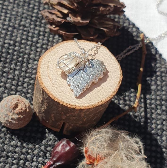 A silver heart shaped leaf pendant hanging on a chain with silver wire wrapped seaglass charm displayed on a tiny log