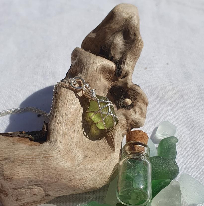 A silver wire wrapped piece of sea glass hanging on a silver chain displayed on a piece of driftwood