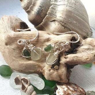 a pair of dangly silver earrings with green and white silver wire wrapped sea glass displayed on a piece of driftwood