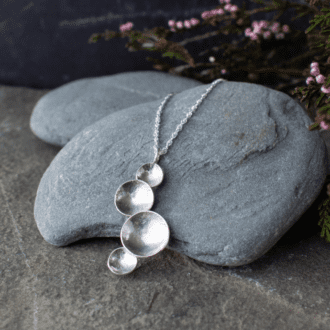 Silver necklace made of four domed circles, resting on a pebble with heather in background