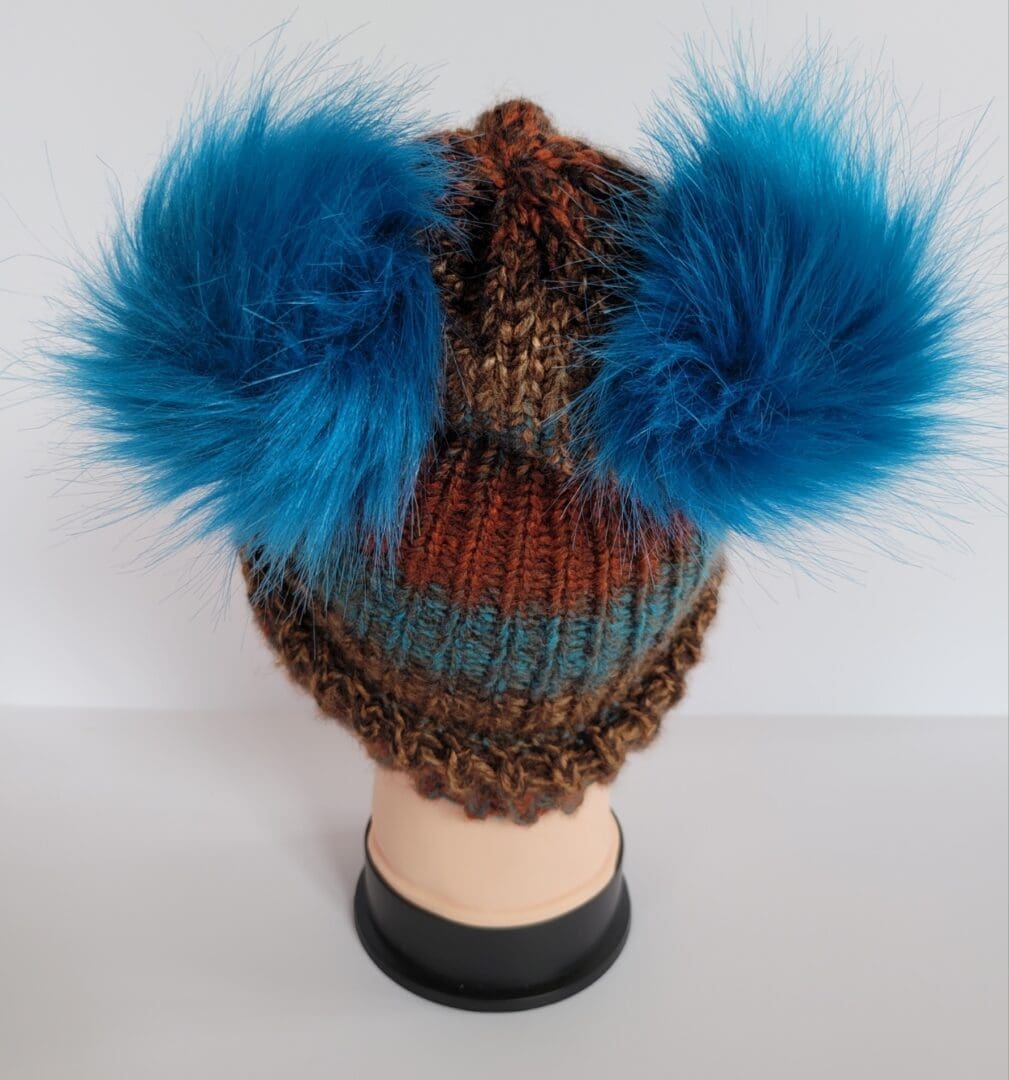 Brown and blue striped hat with brim featuring two blue fluffy pom poms