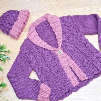 Childs matching handknitted hat and jacket in purple and pink wool with cable stitch detail.